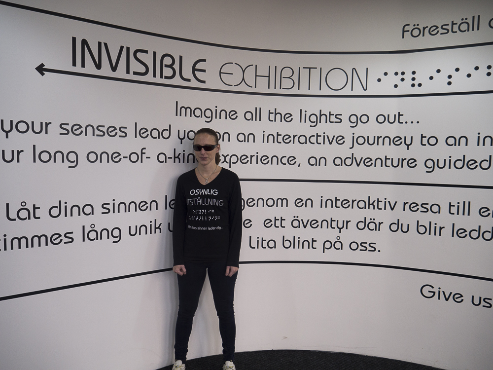 Annika Wistrand Elfving, svartklädd med solglasögon och håret i hästsvans framför utställningsbilden till Osynlig utställning