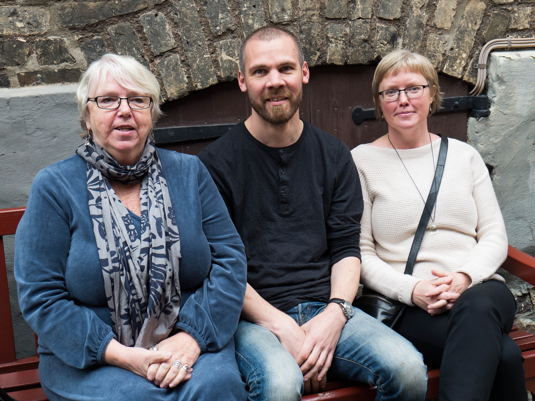 Från vänster sitter Birgitta Lööv, Hugos mormor i vitt kort hår, gasögon och en blå kläning. I mitten pappa Olle med kort skgg, svart tröja och jeans och till höger Malin med ljus tröja. De sitter utomhus på en bänk.