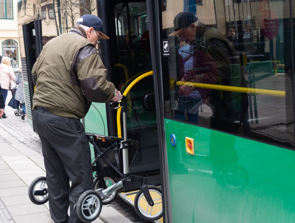 en man i keps lyfter sin rullator ombord på en grön buss