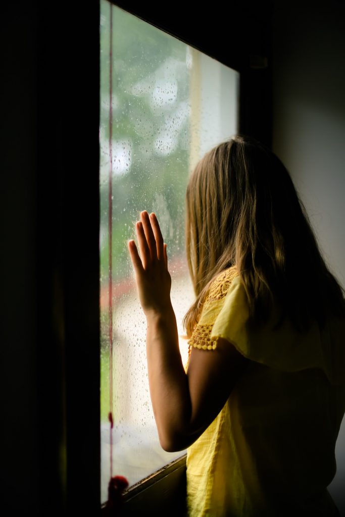 Foto: ung kvinna i gul tröja som tittar ut genom ett fönster