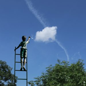 Mörkgrå staty med en pojek som klättrar på en fristående stege för att nå ett moln på en blå himmel. Stegen sträcker sig ovanför några trädtoppar. Foto.